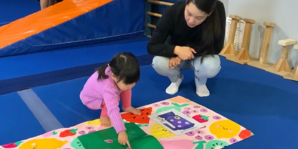 A girl toddler playing on a colorful mat with her coach.